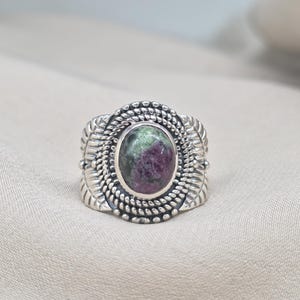 Puede incluir: Anillo de plata con una piedra preciosa ovalada con tonos verdes y morados. El anillo tiene un diseño detallado con un borde trenzado y detalles en forma de hoja. Una pieza de joyería.