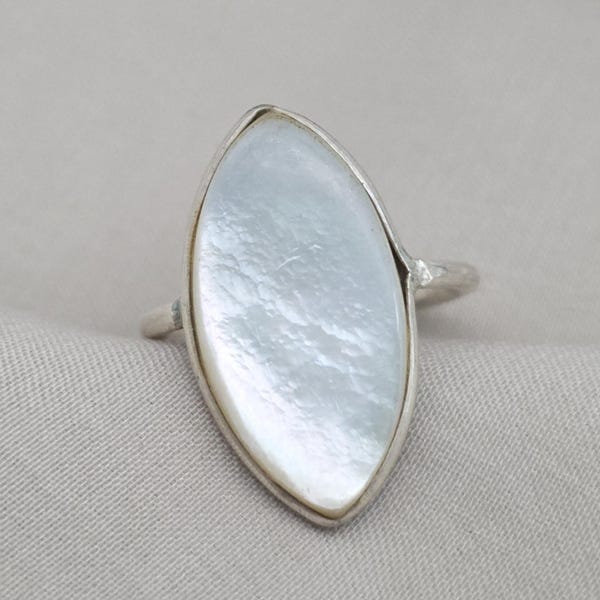 Può includere: Anello in argento con una grande pietra bianca iridescente di forma ovale. La pietra è incastonata in una semplice fascia d'argento, creando un design classico ed elegante. L'anello è esposto su un tessuto neutro.
