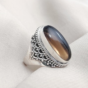 May include: A silver ring featuring an oval gemstone with shades of grey and brown. The ring has detailed silverwork around the stone and on the band. The background is a white fabric.