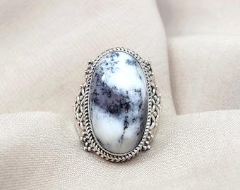Puede incluir: Anillo de plata ovalado con una piedra blanca con inclusiones negras y grises. La piedra está engastada en una banda de plata decorativa con un diseño de filigrana. El anillo es una pieza llamativa con un diseño detallado.