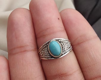 May include: A silver ring with an oval, light blue turquoise stone. The ring has a detailed, braided design around the stone and along the band. The ring is on a finger.