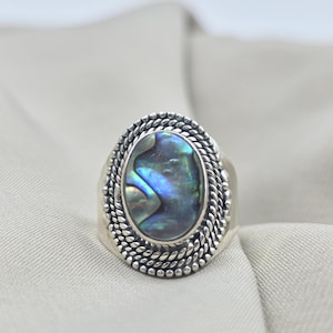 Peut inclure: Bague en argent ornée d'une coquille d'ormeau ovale. La coquille irisée présente des nuances de bleu, de vert et de violet. La bague est bordée d'une bordure texturée en forme de corde.