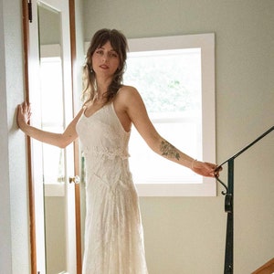 May include: A cream-colored lace wedding dress with a fitted bodice and a flowing skirt. The dress features delicate floral lace detailing and spaghetti straps. The bride is standing on a wooden staircase near a window.