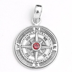 May include: Silver compass pendant with a central red gemstone. The pendant has directional markings (N, S, E, W) and a detailed, textured design. The bail is integrated into the top of the pendant.