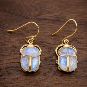 May include: A pair of gold-toned drop earrings featuring a beetle-shaped design. Each earring showcases a smooth, iridescent moonstone cabochon set within a gold metal frame, suspended from a simple curved gold-toned ear wire hook.