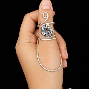 May include: A silver-toned metal ring featuring a spiral design that wraps around the thumb. The ring includes a prominent floral motif with detailed petals and a textured center, connected to a smooth, elongated wire loop extending downward.