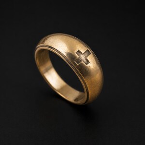 Peut inclure: Une bague dorée avec un motif de croix. La bague a un anneau arrondi et un symbole de croix gravé sur la surface. La bague est présentée sur un fond sombre.