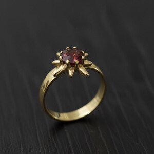 May include: A gold ring featuring a round, deep red gemstone. The gemstone is set in a starburst design, with pointed prongs holding it in place. The ring's band is smooth and polished, creating a classic and elegant look.