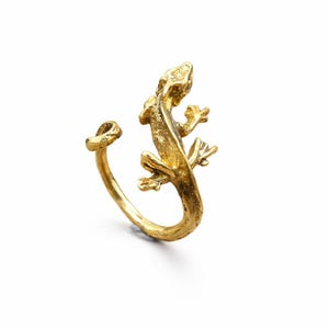 May include: A gold-colored ring in the shape of a lizard. The lizard is detailed with visible legs, a tail, and a head. The ring wraps around the finger with an open design. The ring is set against a white background.