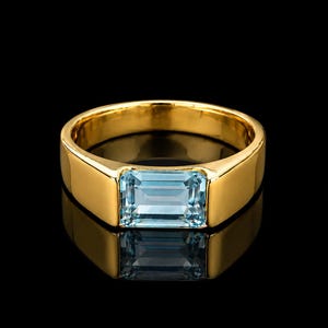 May include: A gold ring featuring a single rectangular emerald-cut light blue gemstone set horizontally in the center. The band is smooth, polished, and solid gold, creating a clean and reflective surface that highlights the clear blue stone.