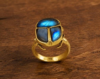 May include: A gold-toned ring featuring a scarab beetle design. The beetle's body is composed of three polished labradorite stones that display iridescent blue flashes. The metal band is smooth and circular, supporting the beetle-shaped setting.