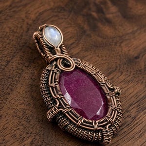 May include: A handcrafted copper wire-wrapped pendant featuring a large, faceted, oval-shaped, deep red gemstone. A smaller, oval, iridescent white gemstone is set at the top. The pendant is displayed on a wooden surface.
