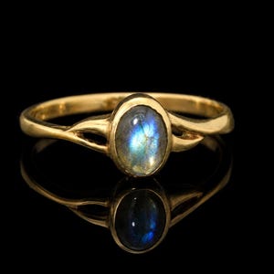 Pode incluir: Um anel de ouro com uma pedra preciosa de labradorite em formato oval e corte cabochão, engastada num bisel liso. A banda apresenta um design dividido em ambos os lados da pedra central, exibindo os reflexos azuis e cinzentos iridescentes.