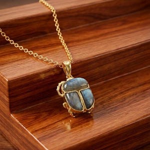 May include: A gold-colored necklace with a pendant shaped like a scarab beetle. The pendant features a blue-grey stone with gold-colored accents. The necklace is displayed on a wooden surface.