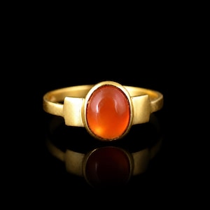 Könnte beinhalten: Ein goldener Ring mit einem glatten, ovalen, orangefarbenen Karneol-Edelstein in der Mitte. Das Band hat ein mattes Finish und ein breites, flaches Design auf beiden Seiten der Steinfassung, was ein klares und ausgewogenes Erscheinungsbild erzeugt.
