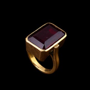 May include: A gold ring featuring a large, rectangular, deep red gemstone set in a thick, polished gold bezel. The stone has a faceted surface that reflects light, and the band is a solid, smooth gold construction with a simple, sturdy design.