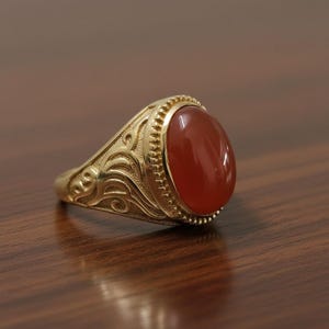 Könnte beinhalten: Ein goldener Ring mit einem ovalen, rot-orangen Edelstein. Der Ring hat ein verziertes, graviertes Design am Band und um den Edelstein. Der Ring liegt auf einer dunkelbraunen Holzoberfläche.