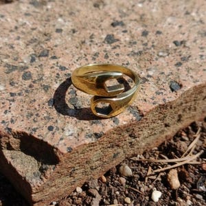 Könnte beinhalten: Ein goldfarbener Ring mit einem schraubenschlüsselartigen Design. Der Ring hat zwei offene Schraubenschlüsselformen, die sich um den Finger wickeln. Der Ring liegt auf einer rauen, rotbraunen Steinoberfläche.
