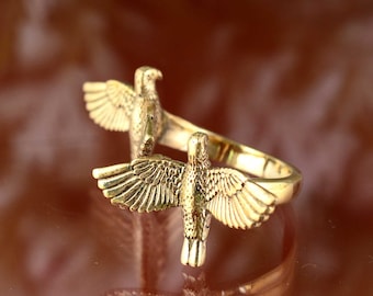 May include: A gold-coloured ring with two detailed eagle designs, each with outstretched wings. The ring has a polished finish and is set against a warm, brown background. The eagles face opposite directions.