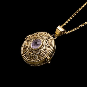 Può includere: Una collana con pendente dorato con un design a medaglione circolare ornato. Il medaglione presenta una pietra preziosa ovale, viola chiaro, al centro. Il pendente è appeso a una delicata catena d'oro, su sfondo nero.