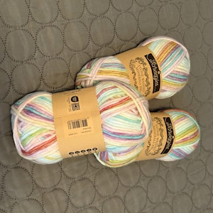 Könnte beinhalten: Drei Knäuel Scheepjes Stone Washed Minerals XXL Garn mit einer bunten Pastellfarbpalette aus Rosa, Gelb, Blau und Weiß. Jedes Knäuel ist mit einer beigen Papierbanderole mit schwarzem Text und einem Strichcode versehen, die den Markennamen zeigt.
