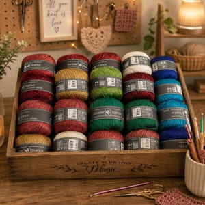 Op de afbeelding: Een houten kist gevuld met kleurrijke garenbollen, waaronder rood, goud, groen, wit en blauw. Het garen is gelabeld met "Starlux" en "Durable". De kist heeft de tekst "CREATE YOUR OWN Magic" op de voorkant. Haaknaalden en scharen zijn ook zichtbaar.