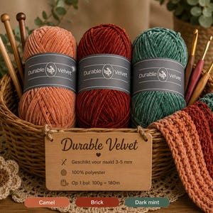 Op de afbeelding: Drie bollen Durable Velvet garen in de kleuren camel, brick en dark mint, elk met een grijs label. Het garen zit in een rieten mand met houten breinaalden, haaknaalden en een houten bordje met productinformatie in het Nederlands.