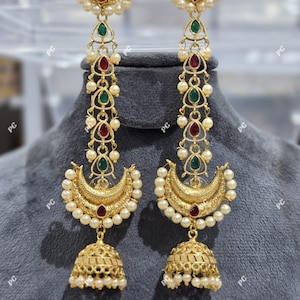 Op de afbeelding: Goudkleurige oorbellen met een halve maan ontwerp, versierd met parels en edelstenen. De oorbellen hebben een klokvormige bodem en worden gepresenteerd op een grijze ondergrond. De edelstenen zijn rood en groen.