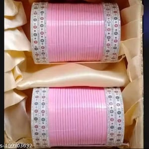 Può includere: Due pile di braccialetti rosa con linee verticali, decorate con fasce bianche con un motivo di piccoli cerchi. I braccialetti sono presentati in una scatola con una fodera satinata.