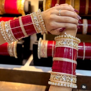 May include: Close-up of red and gold bangles. The bangles are adorned with gold accents and clear, sparkling stones. The design features multiple layers of red and gold, creating a luxurious and ornate appearance. The jewelry is displayed on a person's wrist.