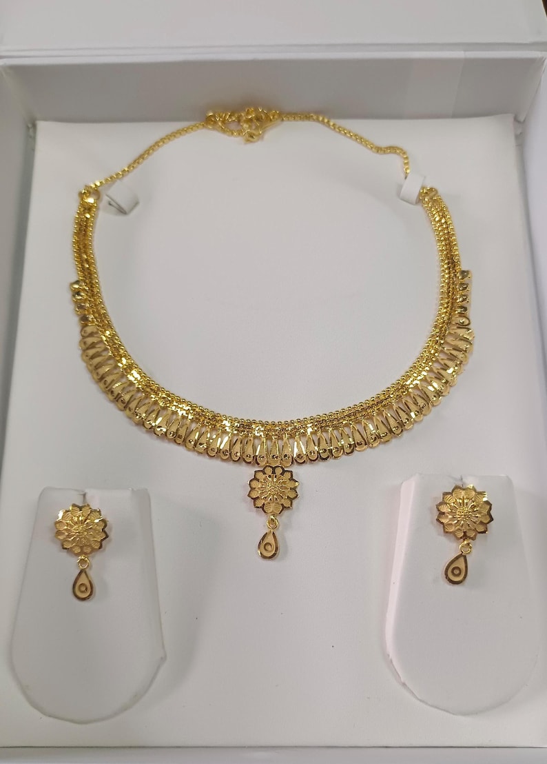Pode incluir: Um conjunto de joias douradas com um colar e brincos a condizer. O colar apresenta um design floral com um pendente central. Os brincos tamb&eacute;m t&ecirc;m um design floral com um pendente em forma de gota. O conjunto &eacute; exibido numa superf&iacute;cie branca.