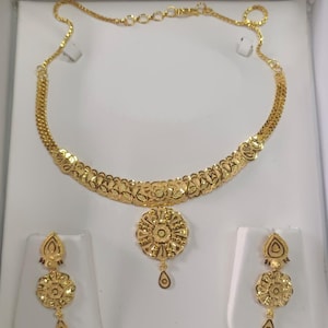Pode incluir: Um conjunto de joias douradas com um colar e brincos a condizer. O colar tem um design floral com um pendente central. Os brincos tamb&eacute;m t&ecirc;m um design floral com um pendente em forma de gota. O conjunto &eacute; exibido sobre um fundo branco.