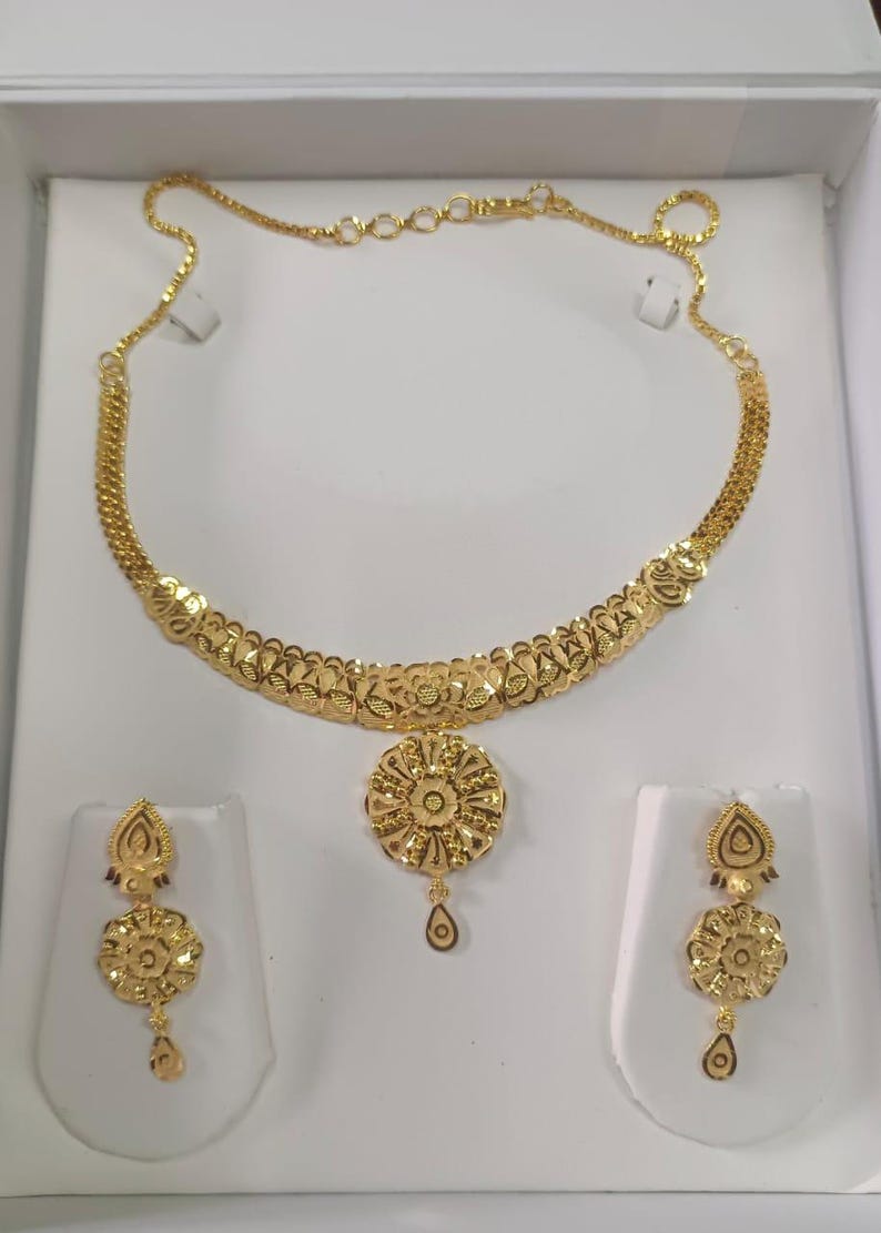 Pode incluir: Um conjunto de joias douradas com um colar e brincos a condizer. O colar tem um design floral com um pendente central. Os brincos tamb&eacute;m t&ecirc;m um design floral com um pendente em forma de gota. O conjunto &eacute; exibido sobre um fundo branco.
