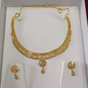 Pode incluir: Um conjunto de colar e brincos dourados. O colar apresenta um pendente floral com um pingente em forma de gota. Os brincos a condizer tamb&eacute;m t&ecirc;m um design floral com um pingente em forma de gota. As joias s&atilde;o exibidas numa caixa branca.