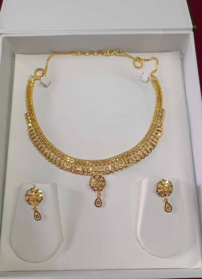 Pode incluir: Um conjunto de colar e brincos dourados. O colar apresenta um pendente floral com um pingente em forma de gota. Os brincos a condizer tamb&eacute;m t&ecirc;m um design floral com um pingente em forma de gota. As joias s&atilde;o exibidas numa caixa branca.