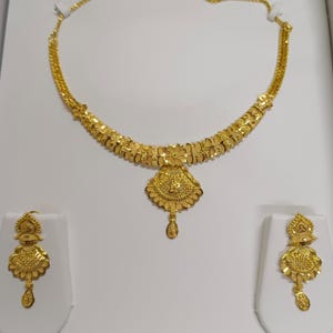 Pode incluir: Um conjunto de joias douradas com colar e brincos a condizer. O colar apresenta um design floral com um pendente central. Os brincos espelham a forma do pendente, criando um visual coeso. O conjunto est&aacute; exposto.