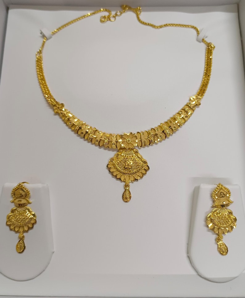 Pode incluir: Um conjunto de joias douradas com colar e brincos a condizer. O colar apresenta um design floral com um pendente central. Os brincos espelham a forma do pendente, criando um visual coeso. O conjunto est&aacute; exposto.