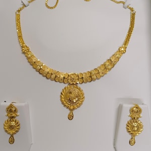 Pode incluir: Um conjunto de colar e brincos dourados. O colar apresenta um design floral com um pendente central. Os brincos espelham o design do pendente. As joias s&atilde;o exibidas em suportes brancos sobre um fundo branco.