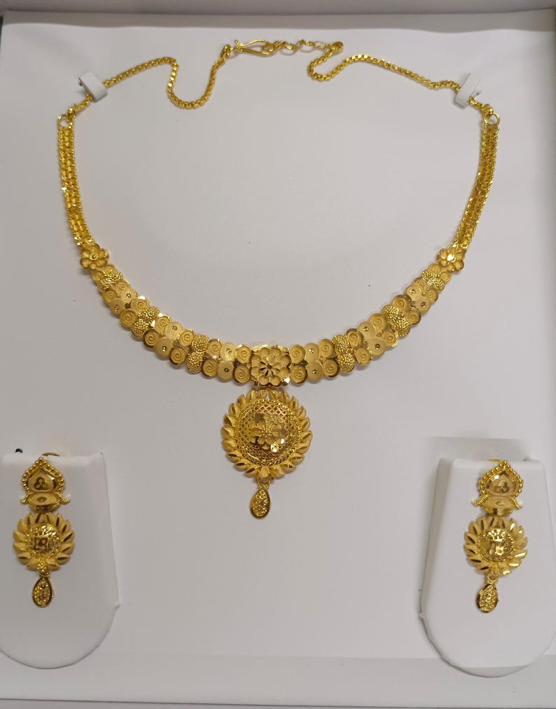 Pode incluir: Um conjunto de colar e brincos dourados. O colar apresenta um design floral com um pendente central. Os brincos espelham o design do pendente. As joias s&atilde;o exibidas em suportes brancos sobre um fundo branco.