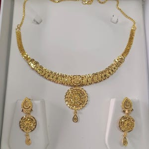 Pode incluir: Um conjunto de joias douradas, incluindo um colar e brincos a condizer, exibido numa caixa branca. O colar apresenta um design floral, e os brincos t&ecirc;m pingentes em forma de gota. O conjunto &eacute; provavelmente feito de ouro ou metal dourado.