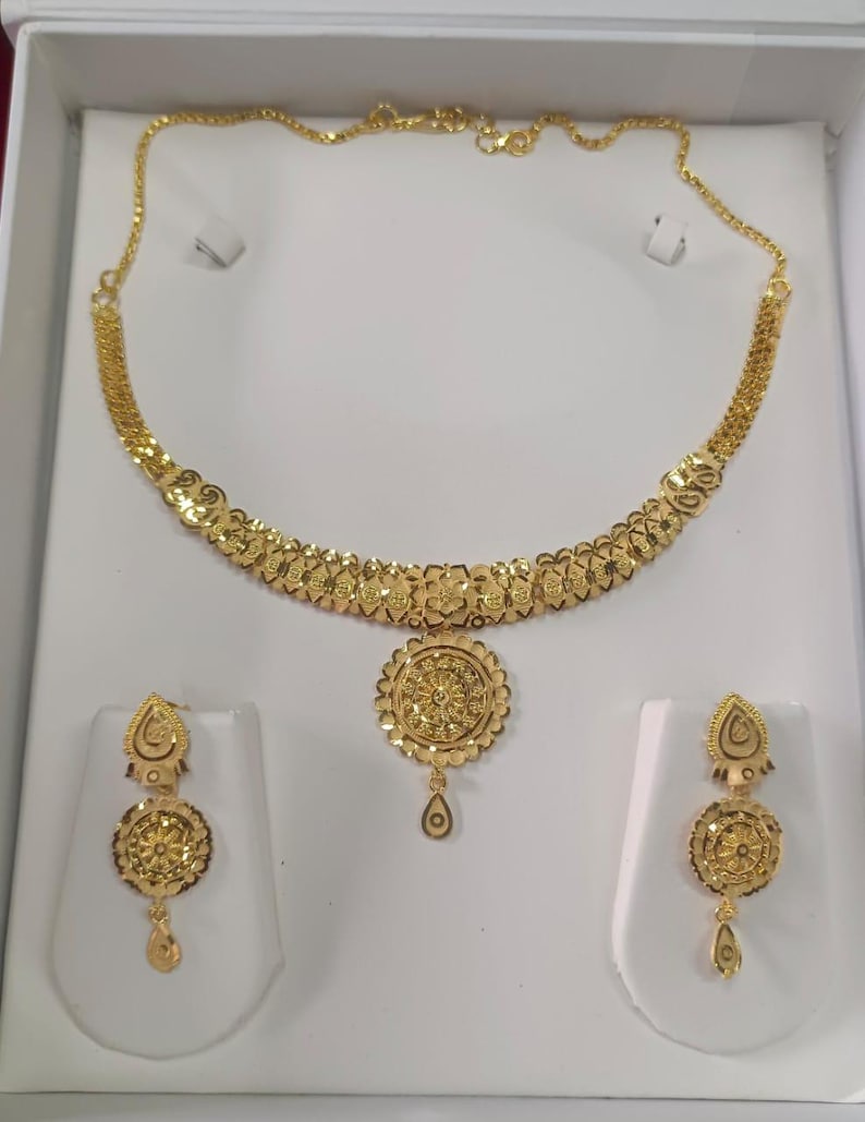 Pode incluir: Um conjunto de joias douradas, incluindo um colar e brincos a condizer, exibido numa caixa branca. O colar apresenta um design floral, e os brincos t&ecirc;m pingentes em forma de gota. O conjunto &eacute; provavelmente feito de ouro ou metal dourado.
