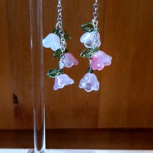 May include: Dangling earrings with pink and white flower charms and green leaf details. The earrings hang from silver-coloured hooks and chains. The floral design is displayed on a clear acrylic stand.
