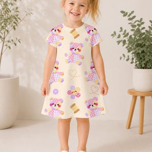 K&ouml;nnte beinhalten: Ein cremefarbenes Kurzarmkleid mit einem sich wiederholenden Muster aus Patchwork-Teddyb&auml;ren in Rosa-, Lila- und Gelbt&ouml;nen, durchsetzt mit kleinen braunen Garnrollen, lila Kn&ouml;pfen und gestrichelten Herzumrissen.
