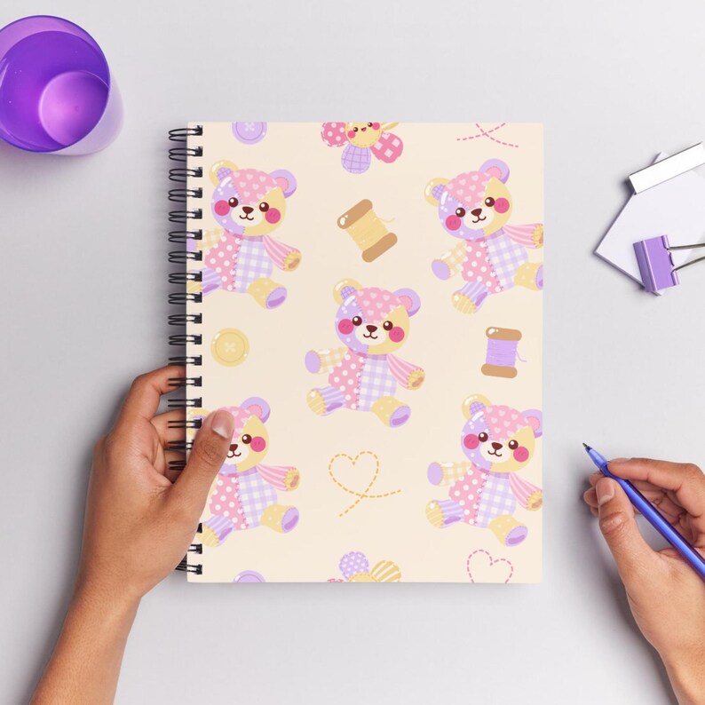 K&ouml;nnte beinhalten: Ein spiralgebundenes Notizbuch mit cremefarbenem Einband, das ein wiederkehrendes Muster aus Patchwork-Teddyb&auml;ren in Rosa-, Lila- und Gelbt&ouml;nen zeigt, durchsetzt mit kleinen Illustrationen von N&auml;hkn&ouml;pfen, Garnrollen und gestrichelten Herzumrissen.