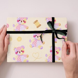 K&ouml;nnte beinhalten: Eine rechteckige Geschenkbox, eingewickelt in cremefarbenes Papier mit einem wiederkehrenden Muster aus Patchwork-Teddyb&auml;ren in Pastellrosa, Lila und Gelb, erg&auml;nzt durch Garnrollen, Kn&ouml;pfe und gestrichelte Herzen, gebunden mit einem dunkelgr&uuml;nen Samtband.