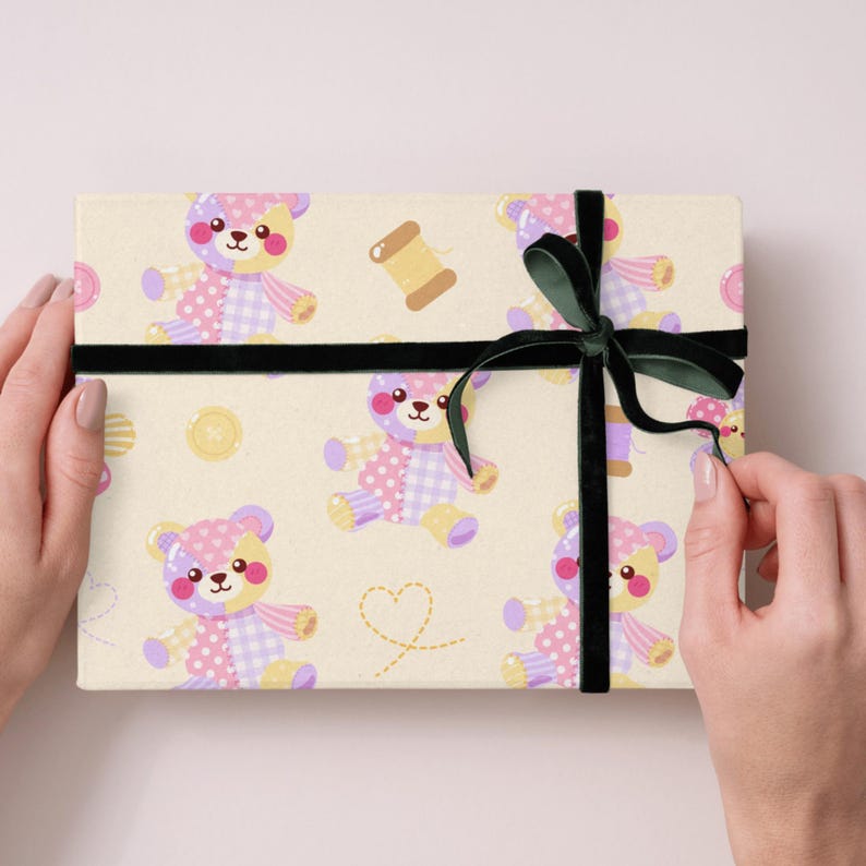 K&ouml;nnte beinhalten: Eine rechteckige Geschenkbox, eingewickelt in cremefarbenes Papier mit einem wiederkehrenden Muster aus Patchwork-Teddyb&auml;ren in Pastellrosa, Lila und Gelb, erg&auml;nzt durch Garnrollen, Kn&ouml;pfe und gestrichelte Herzen, gebunden mit einem dunkelgr&uuml;nen Samtband.