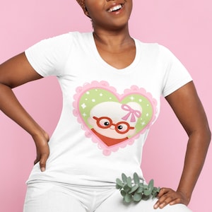 May include: White v-neck t-shirt featuring a heart-shaped graphic with a cartoon image of a woman with glasses, a red scarf, and a pink bow. The heart has a pink scalloped border and a light green interior with white stars.