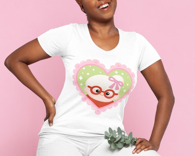 May include: White v-neck t-shirt featuring a heart-shaped graphic with a cartoon image of a woman with glasses, a red scarf, and a pink bow. The heart has a pink scalloped border and a light green interior with white stars.