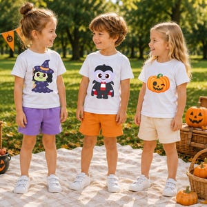 May include: Three children wearing white Halloween-themed t-shirts and shorts. The shirts feature a witch, a vampire, and a jack-o'-lantern. The shorts are purple, orange, and beige. The children are standing on a blanket outdoors.