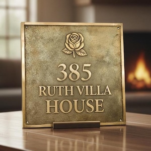 Op de afbeelding: Een rechthoekig messing huisnummerbord met het nummer 385, de woorden "RUTH VILLA HOUSE" en een rozenembleem. Het bord staat op een houten tafel. De achtergrond is wazig, met een open haard zichtbaar.